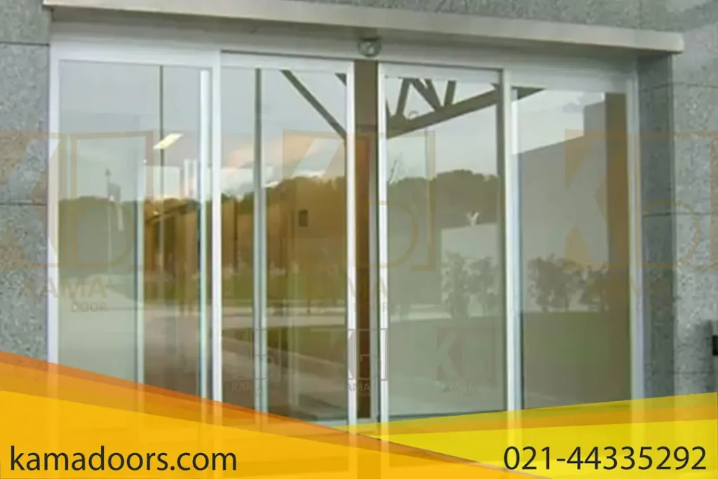 مجموعه ای از درب های شیشه ای اتوماتیک دو لت را در ورودی ساختمان نشان می دهد. درب های اتوماتیک کامادر، دارای قاب و دستگیره فلزی هستند که انعکاس آن بر روی سطح شیشه قابل مشاهده است. بالای درب ها، یک لنگه سنگی وجود دارد و دیوار بیرونی ساختمان به نظر می رسد که از سنگ یا بتون ساخته شده باشد. یک فرش مشکی در جلوی درب ها قرار داده شده است. انعکاس روی شیشه یک محیط بیرونی با درختان و آسمان را نشان می دهد که نشان می دهد عکس در طول روز گرفته شده است.