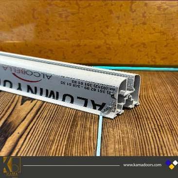 دو قطعه آلومینیومی سفید رنگ است که به یکیدیگر متصل شده و به عنوان یراق آلات شیشه های بالکنی مورد استفاده قرار می گیرد. این قطعه آلومینیومی بر روی یک میز چوبی با رگه های مشکی قرار گرفته است.