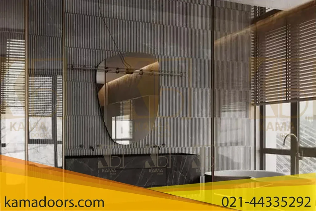 یک فضای داخلی مدرن، احتمالاً حمام، با طراحی معاصر با پارتیشن‌ های شیشه‌ ای و پرداخت‌ های پیشرفته را به تصویر می‌ کشد. پارتیشن های شیشه ای دارای بافت آجدار هستند که هم حریم خصوصی و هم شفافیت را فراهم می کنند. در مرکز تصویر، یک آینه بیضی شکل با یک قاب فلزی روی پارتیشن شیشه ای نصب شده است. زیر آینه، یک میز سنگ مرمر تیره یا سنگ وجود دارد که به زیبایی مدرن فضا می افزاید. دکور کلی طرحی مینیمال و مدرن را پیشنهاد می کند، احتمالاً برای یک فضای اداری یا مسکونی. وجود نور طبیعی که از سمت راست تصویر، احتمالاً از طریق پنجره ای با منظره ای از فضای سبز وارد می شود، یک محیط آرام و طبیعی به فضا می بخشد. نورپردازی ملایم و محیطی به نظر می رسد و به جو کلی کمک می کند.