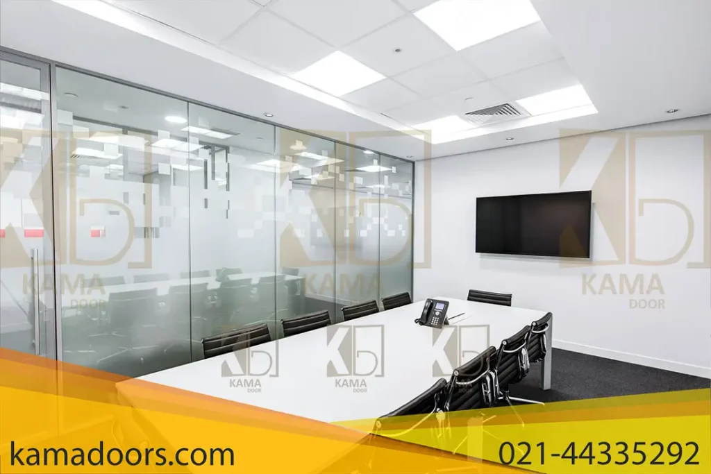نشان دهنده یک اتاق کنفرانس مدرن است. اتاق دارای یک میز کنفرانس بزرگ سفید با صندلی‌ های اداری سیاه است. در یک طرف اتاق، یک تلویزیون صفحه تخت دیواری وجود دارد و در وسط میز، به نظر می‌ رسد یک تلفن کنفرانس قرار دارد. اتاق توسط پارتیشن های شیشه‌ ای کامادر با الگو های مات برای حفظ حریم خصوصی احاطه شده است و برچسب‌ های مستطیلی به رنگ قرمز و خاکستری روی شیشه وجود دارد. کف اتاق با فرش خاکستری پوشیده شده و سقف دارای نورپردازی توکار است که فضا را به طور یکنواخت روشن می‌ کند.