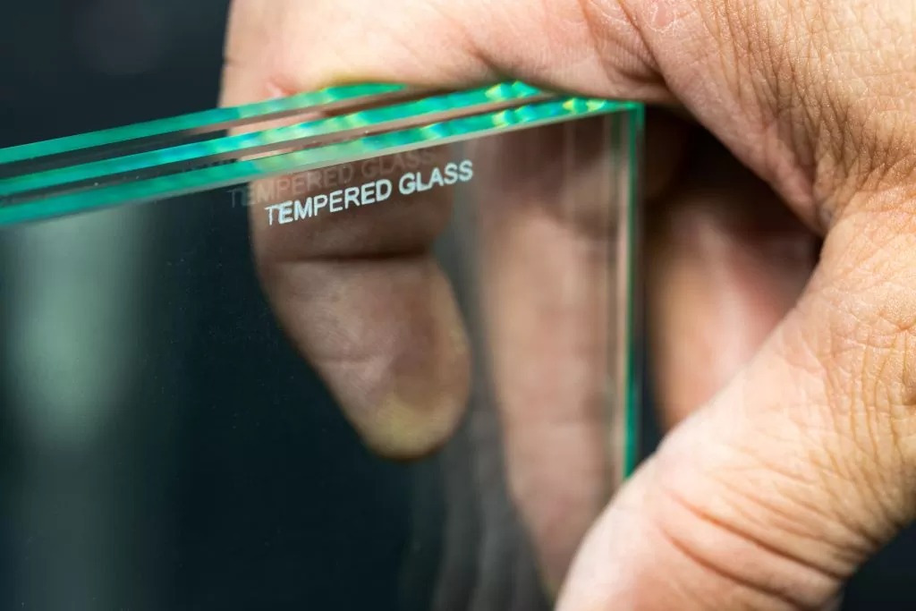 نمای نزدیک از دست شخصی دیده می شود که پشته‌ای از صفحات شیشه‌ای شفاف را در دست گرفته است. پانل های شیشه ای شفاف هستند و عبارت "TEMPERED GLASS" به طور مکرر در یک لبه با فونت سفید چاپ شده است که نشان دهنده نوع شیشه و استحکام بیشتر آن در مقایسه با شیشه معمولی است. لبه هایی که پانل ها در تماس با انگشتان هستند برجسته می شوند، احتمالاً به دلیل فشار اعمال شده توسط دست یا خاصیت انکساری جزئی لبه های شیشه. تمرکز روی انگشتان است که روی شیشه فشار می‌آورند و برجستگی‌ها و بافت‌های پوست را آشکار می‌کنند. پس زمینه تار است و توجه را به وضوح و کیفیت پانل های شیشه ای معطوف می کند.