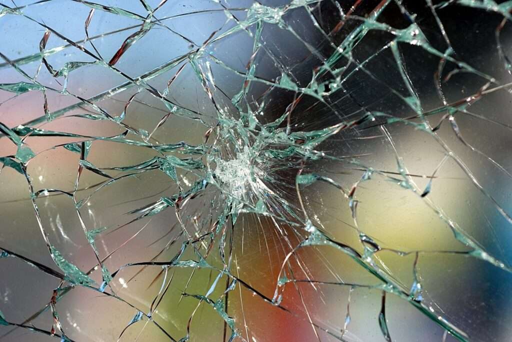 نمای نزدیکی از شیشه های شکسته سکوریت را نشان می دهد که الگوی پیچیده ای از شکستگی را با نقطه برخورد مرکزی و ترک های تابشی به تصویر کشیده است. شیشه سکوریت به شدت تکه تکه شده است، با قطعات بزرگتر و ناهموار و شبکه ای از خطوط که در سراسر سطح پخش شده است. پس زمینه از میان شیشه سکوریت شکسته تار به نظر می رسد، که نشان می دهد زمانی بخشی از یک سازه، احتمالاً یک پنجره یا درب بوده است. رنگ‌ های مختلف در پس‌ زمینه نشان‌دهنده انعکاس یا نمایی از سمت دیگر است.
