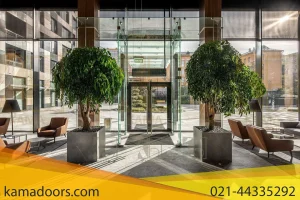 یک لابی ساختمان مدرن را با نمای واضح از میان درب های شیشه ای به حیاط بیرونی نشان می دهد. لابی متقارن است و دارای دو گیاه گلدانی بزرگ در دو طرف ورودی، و چهار صندلی قهوه‌ ای رنگ با میز های کناری مربعی در بین آن‌ ها است. به نظر می رسد که کفپوش سنگ یا کاشی صیقلی است که نور طبیعی را که از درب اتوماتیک کامادر به فضا سرازیر می شود، منعکس می کند. حیاط بیرون دارای ساختمان های اضافی و درختان در پس زمینه است.