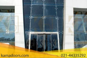 نمای روبروی یک ساختمان مدرن با ورودی شیشه ای بزرگ را نشان می دهد. نمای شیشه ای کامادر متشکل از پانل های شیشه ای بلند و عمودی است که در یک قاب خاکستری قرار گرفته اند و در دو طرف با دیوارهای محکم احاطه شده است. مجموعه ای از پله های بتنی به ورودی منتهی می شود که نشان می دهد این نقطه دسترسی اصلی به ساختمان است. آسمان صاف و آبی است که نشان دهنده شرایط آب و هوایی مناسب است. انعکاس درختان برهنه در شیشه دیده می شود که عنصری از محیط اطراف را به نمای ساختمان اضافه می کند.
